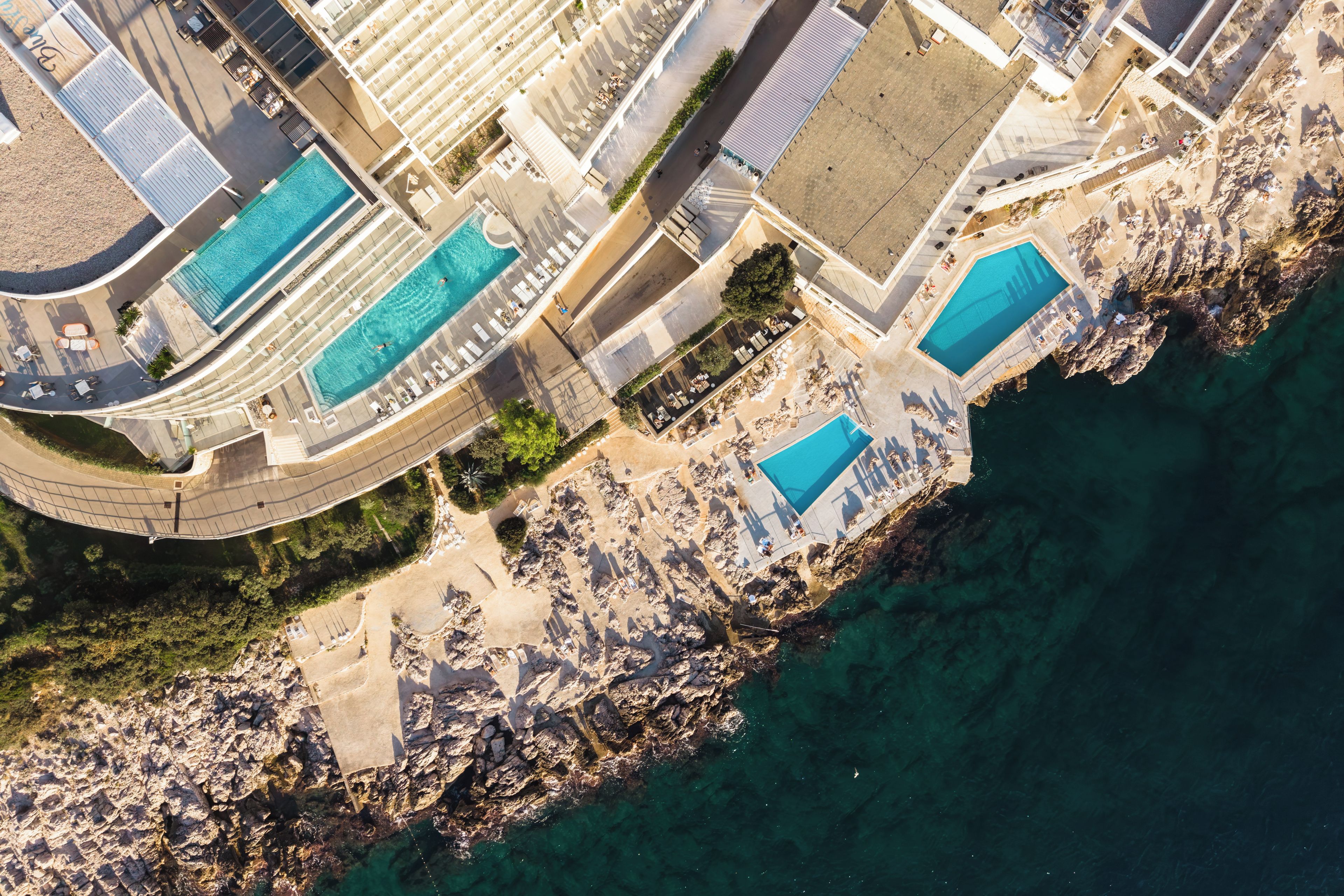 An aerial view of a coastal hotel with multiple swimming pools, a rocky shoreline, and a paved walkway.