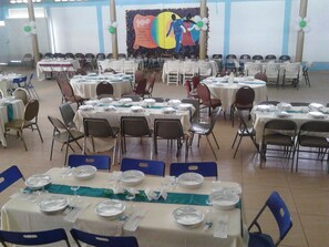 Une salle de banquet avec de nombreuses tables rondes dressées pour le repas, des chaises disposées autour, et une scène avec un décor coloré en arrière-plan.