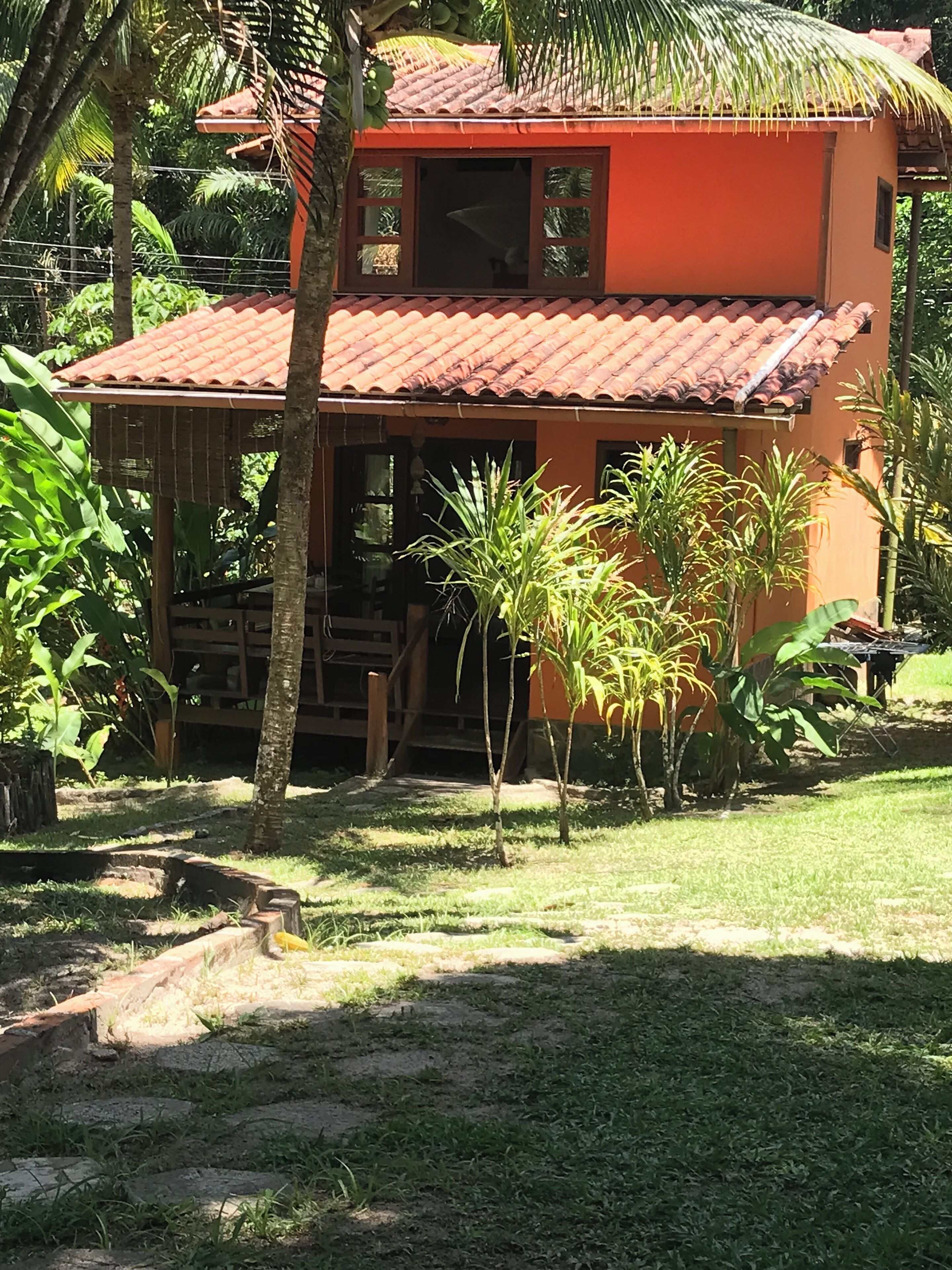 Uma casa de dois andares com telhado de telhas vermelhas, cercada por vegetação exuberante e palmeiras.