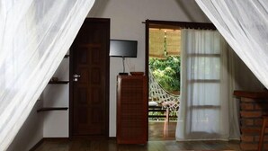 A bedroom with a bed, a TV, a wooden cabinet, a door, and a view of a hammock and greenery outside. - Vila Sofia chalès (Cairu)