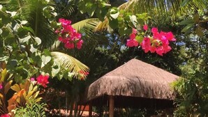 A thatched-roof structure surrounded by lush greenery and pink flowers. - Vila Sofia chalès (Cairu)