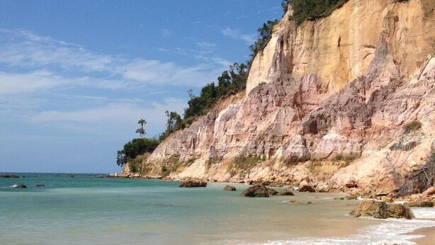 A beach with clear turquoise water, a sandy shore, and a rocky cliff with sparse vegetation. - Vila Sofia chalès (Cairu)