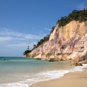 A beach with clear turquoise water, a sandy shore, and a rocky cliff with sparse vegetation. - Vila Sofia chalès (Cairu)