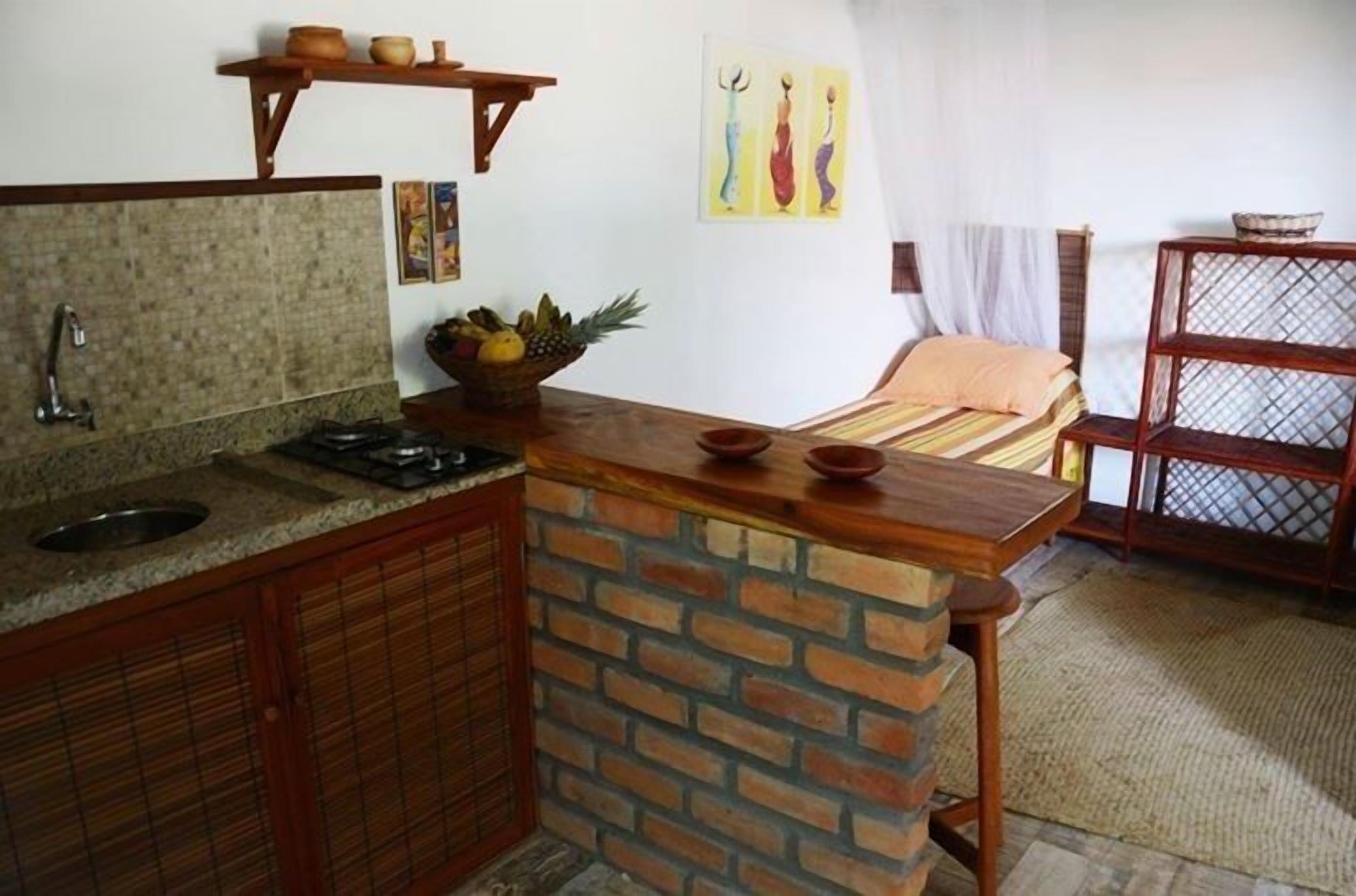 A compact kitchen with a granite countertop, a brick backsplash, and wooden cabinets. There is a small sink, a stove, and a fruit bowl on the counter.