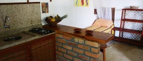 A compact kitchen with a granite countertop, a brick backsplash, and wooden cabinets. There is a small sink, a stove, and a fruit bowl on the counter.