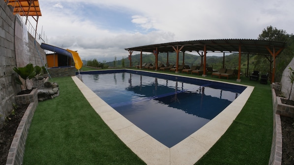 Una piscina con tobogán, rodeada de césped artificial y una zona de estar cubierta.