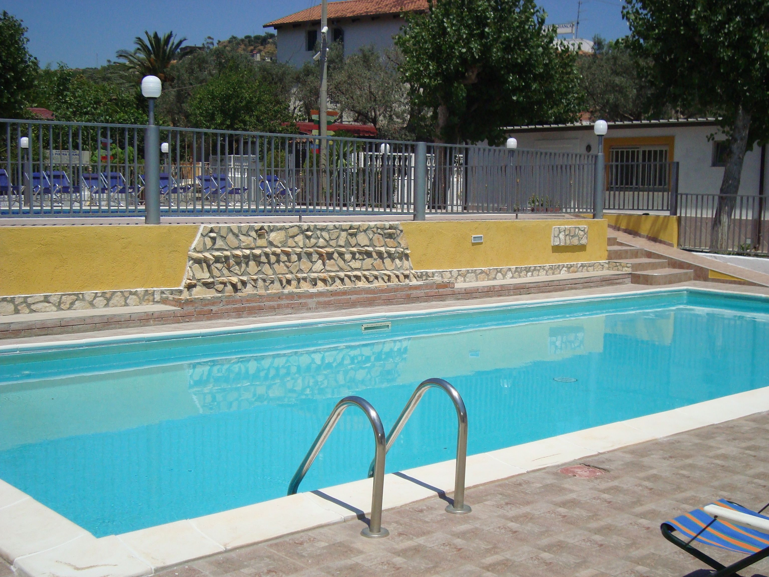 A swimming pool with a stone wall and metal railings, surrounded by a fence and a few lounge chairs.