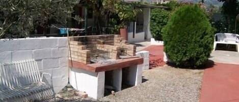 Un jardin avec un espace barbecue, une chaise blanche et un petit arbre.