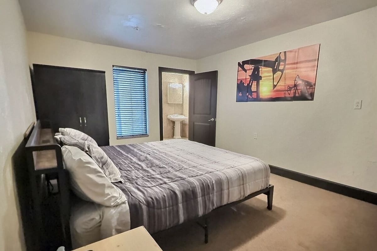Ein Schlafzimmer mit Bett, Schreibtisch, Stuhl, einem Fenster mit Jalousien, einer Tür und einem Wandbild.