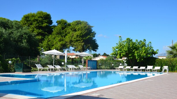 Una piscina con sillas de descanso y sombrillas, rodeada de árboles y un cielo despejado.