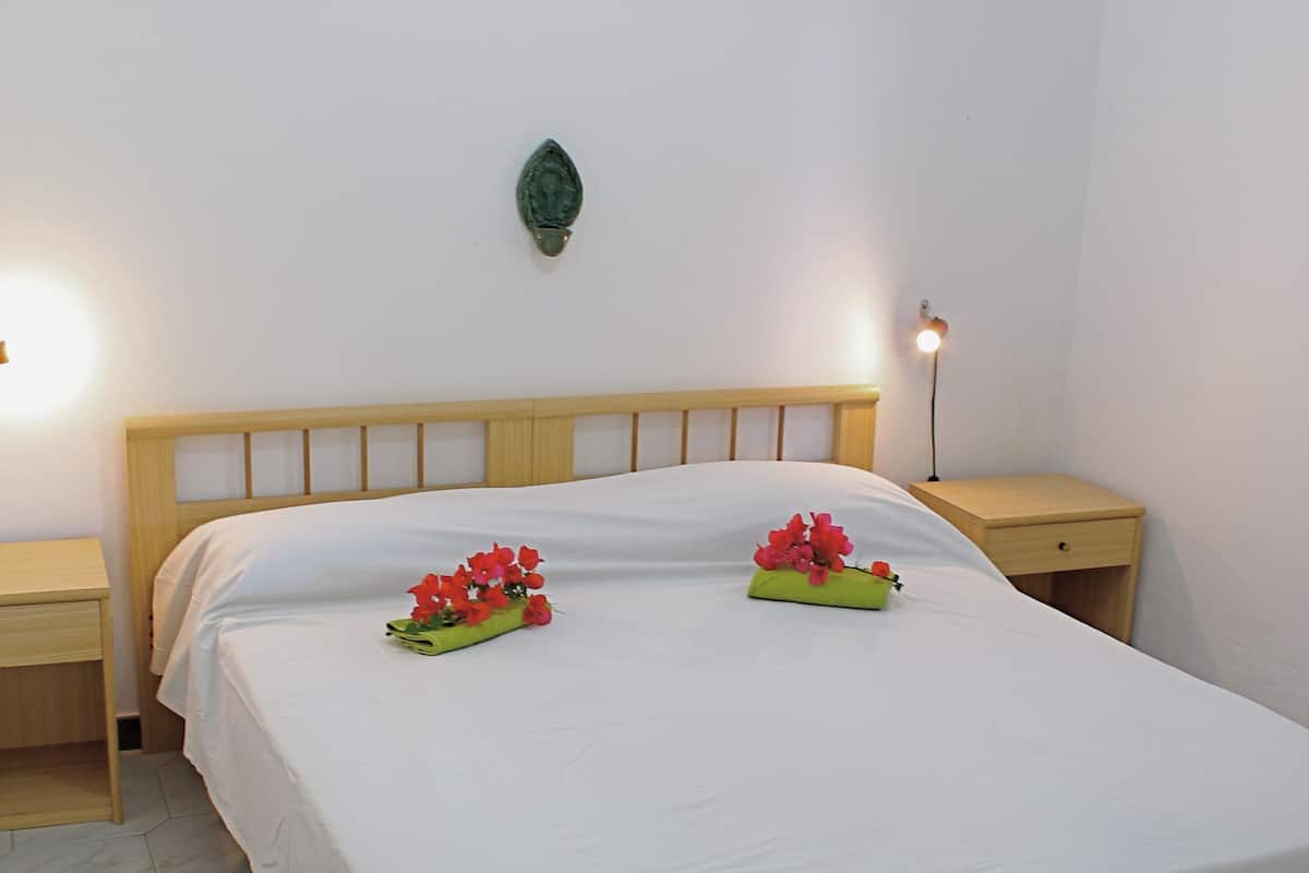 Cama bem arrumada com colcha branca e dois arranjos de flores vermelhas. Duas mesas de cabeceira, uma de cada lado da cama, cada uma com uma luminária. O quarto tem um design simples e clean, com cabeceira de madeira e um pequeno objeto decorativo na parede.