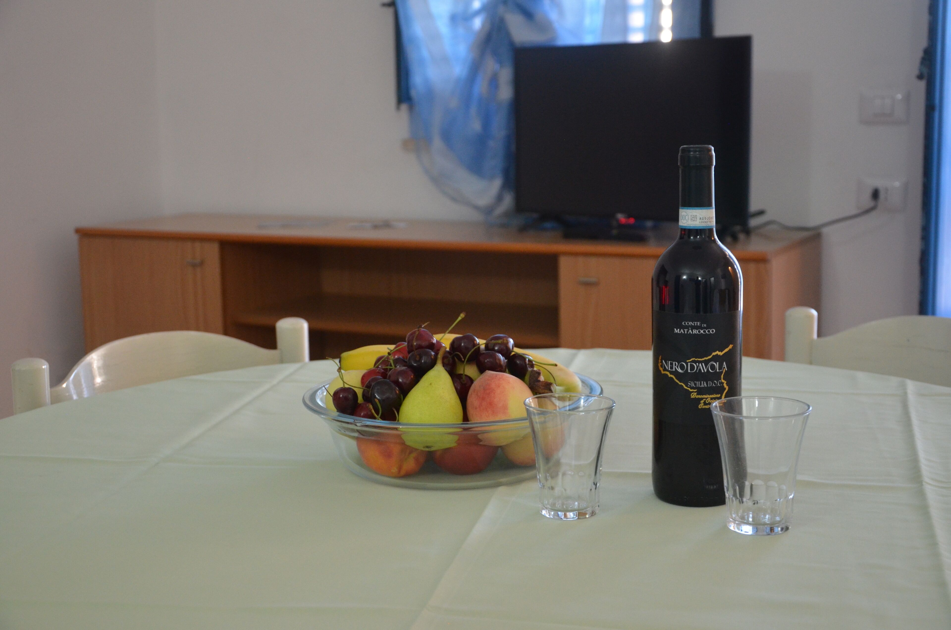 A dining table with a fruit bowl, a bottle of wine, and two shot glasses.