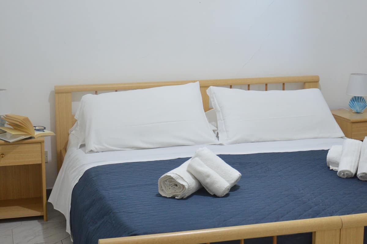 Cama bem arrumada com colcha azul e almofadas brancas, cabeceira de madeira, mesa de cabeceira com abajur e um quadro com uma cena de praia na parede.