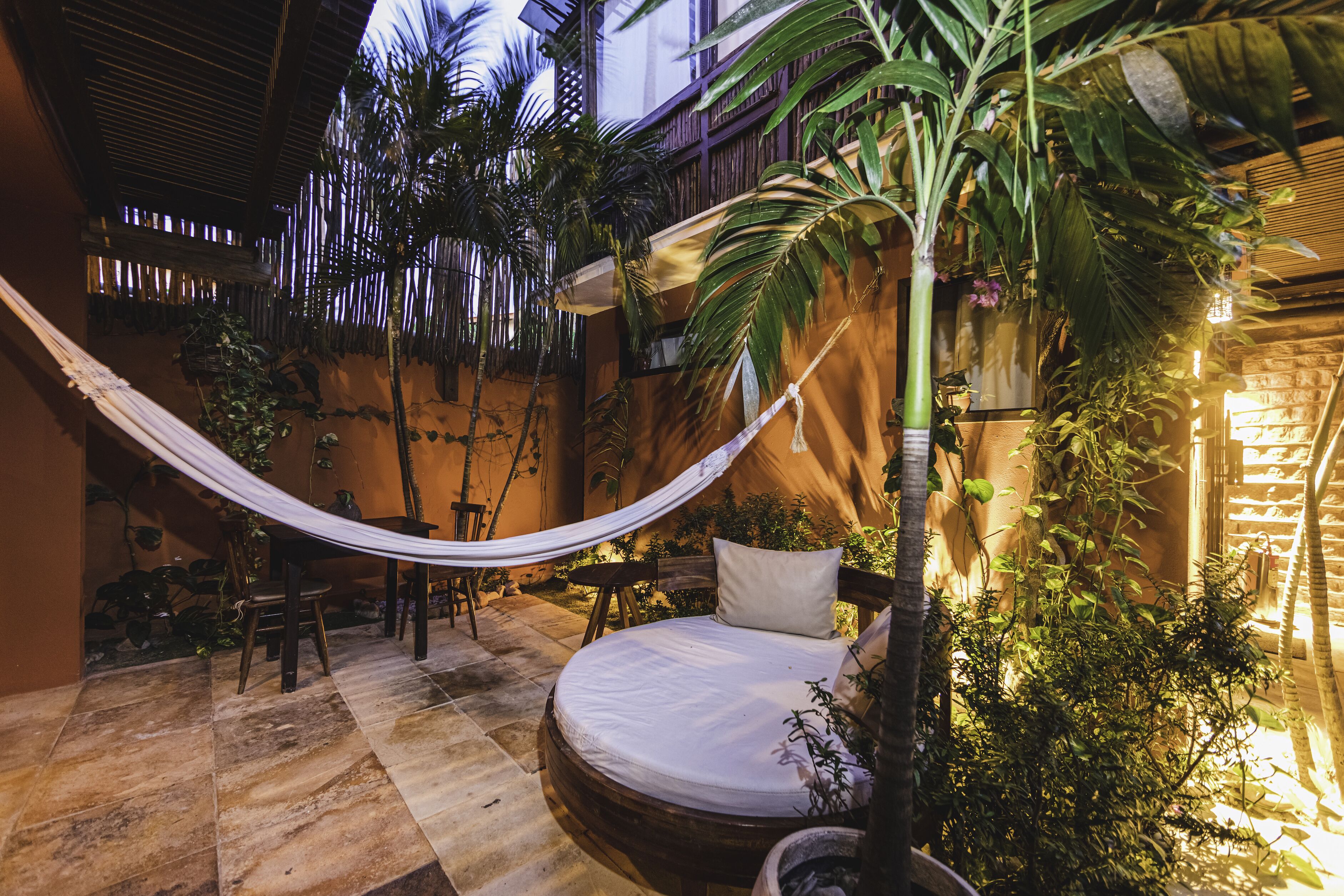 A hammock, a round bed, and potted plants in a courtyard with a wooden fence and a thatched roof.