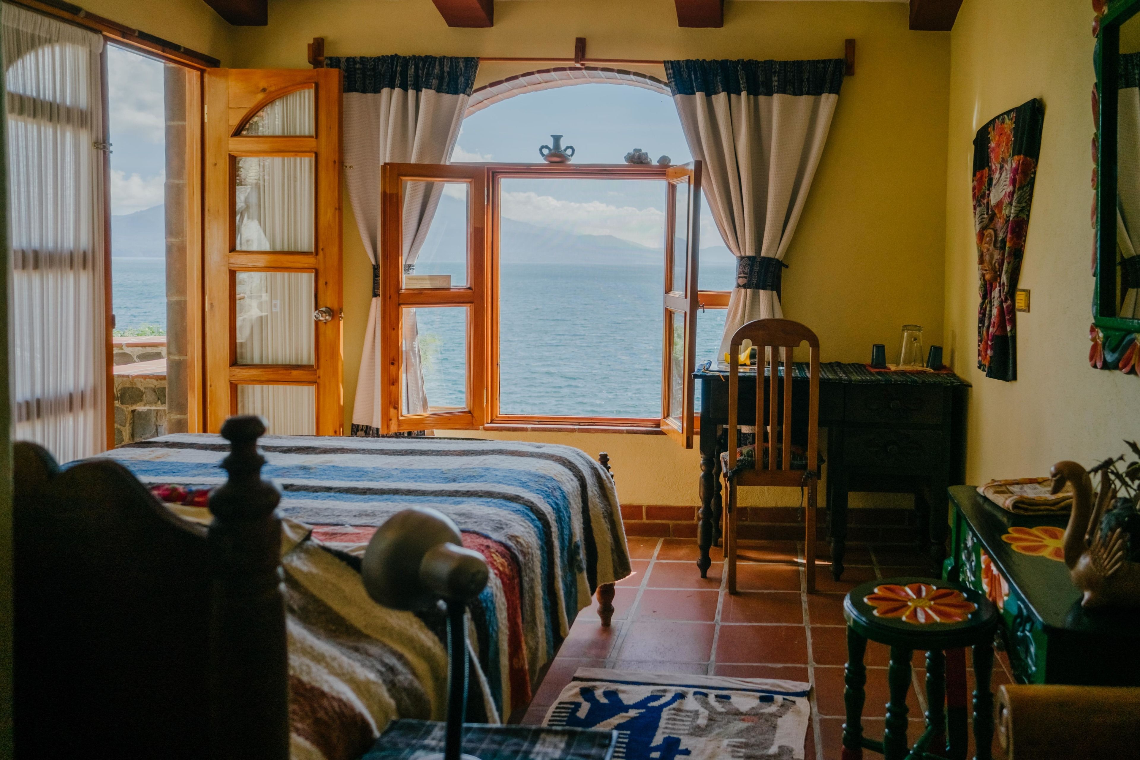 Ein Zimmer mit einem Bett, einem hölzernen Stuhl, einem kleinen Tisch und Blick auf das Meer durch eine offene Tür.