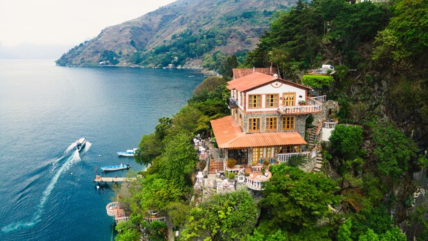 A large, multi-story house with a red roof, situated on a cliff overlooking a body of water. - Hotel y Cafe La Casa del Mundo (Santa Cruz La Laguna)