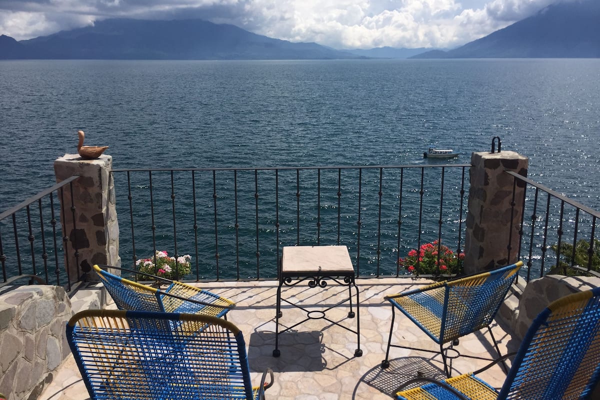 A terrace with a view of a lake and mountains, featuring stone pillars, a table, and blue chairs.