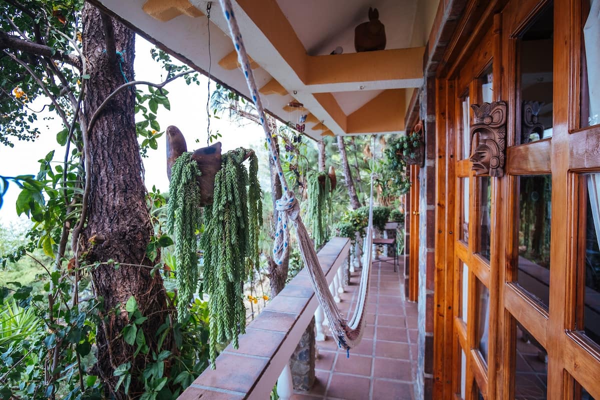 A wooden balcony with hanging plants, a hammock, and a stone bench.
