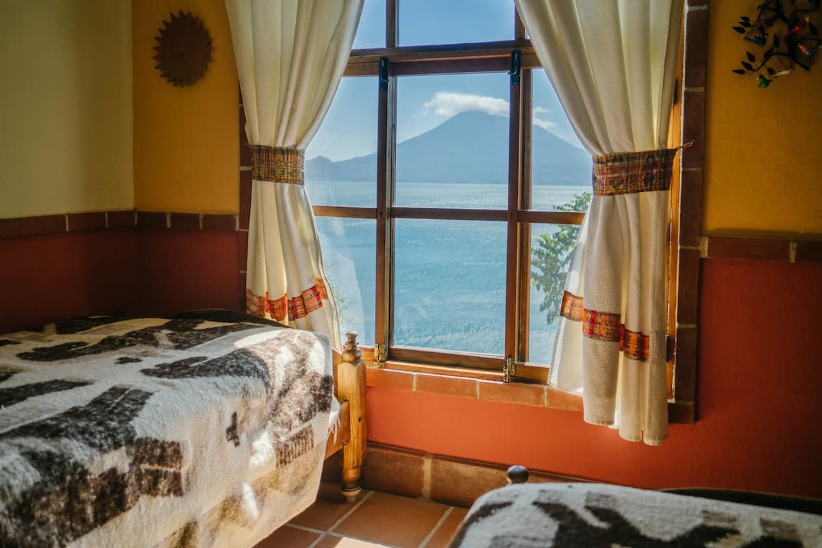 A room with two beds, a window with curtains, a bottle on the floor, and a view of the sea and a mountain.