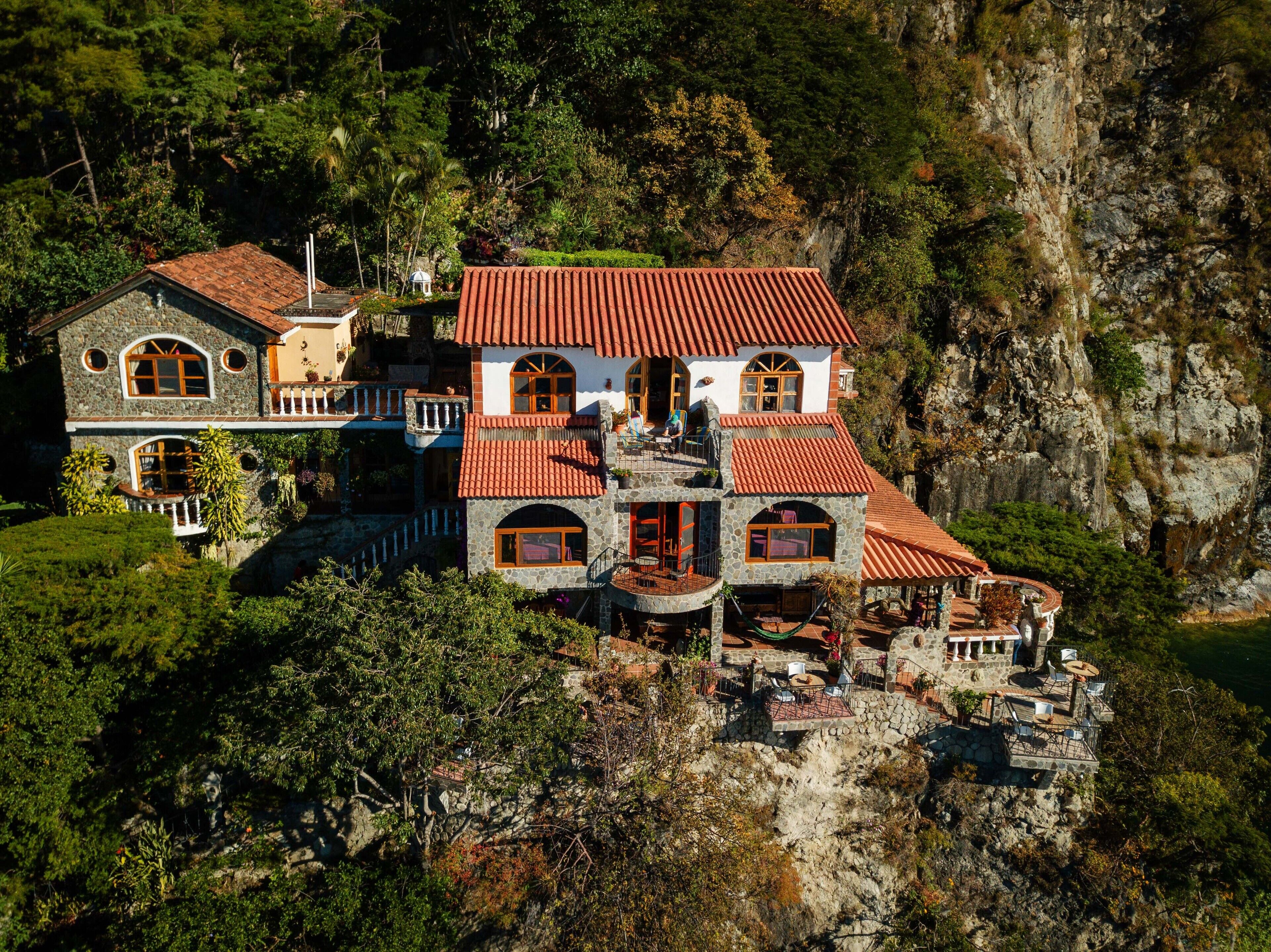 A large, multi-story stone house with red-tiled roofs, situated on a cliff with lush greenery and rocky terrain.