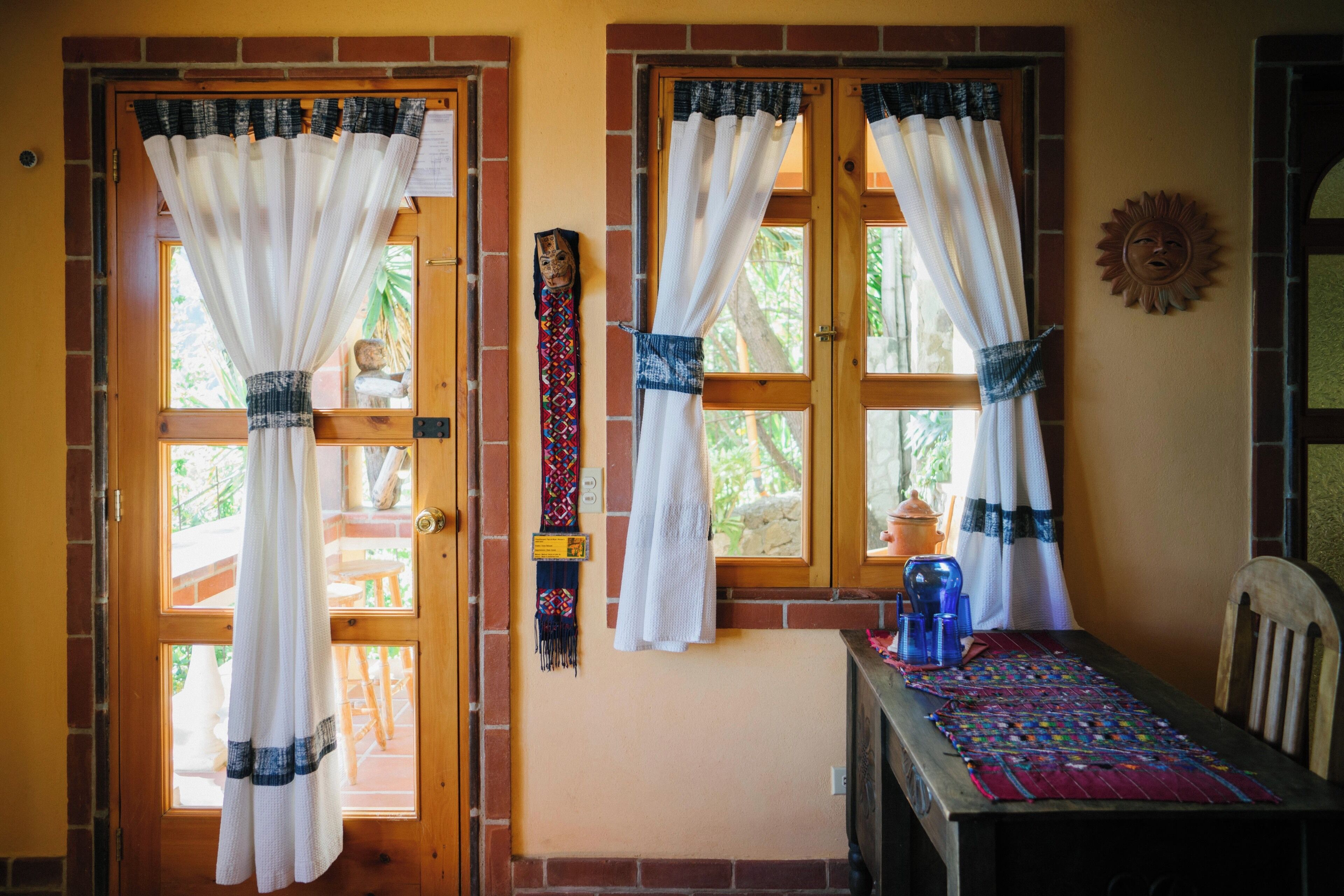 A room with two wooden doors with white curtains, a small table with a colorful tablecloth, and a chair.