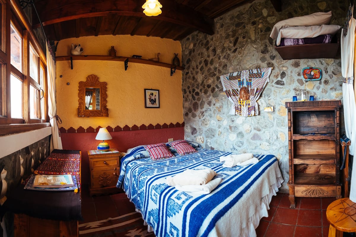 A cozy room with a bed, a wooden nightstand, a small table, a mirror, and a shelf with decorative items.