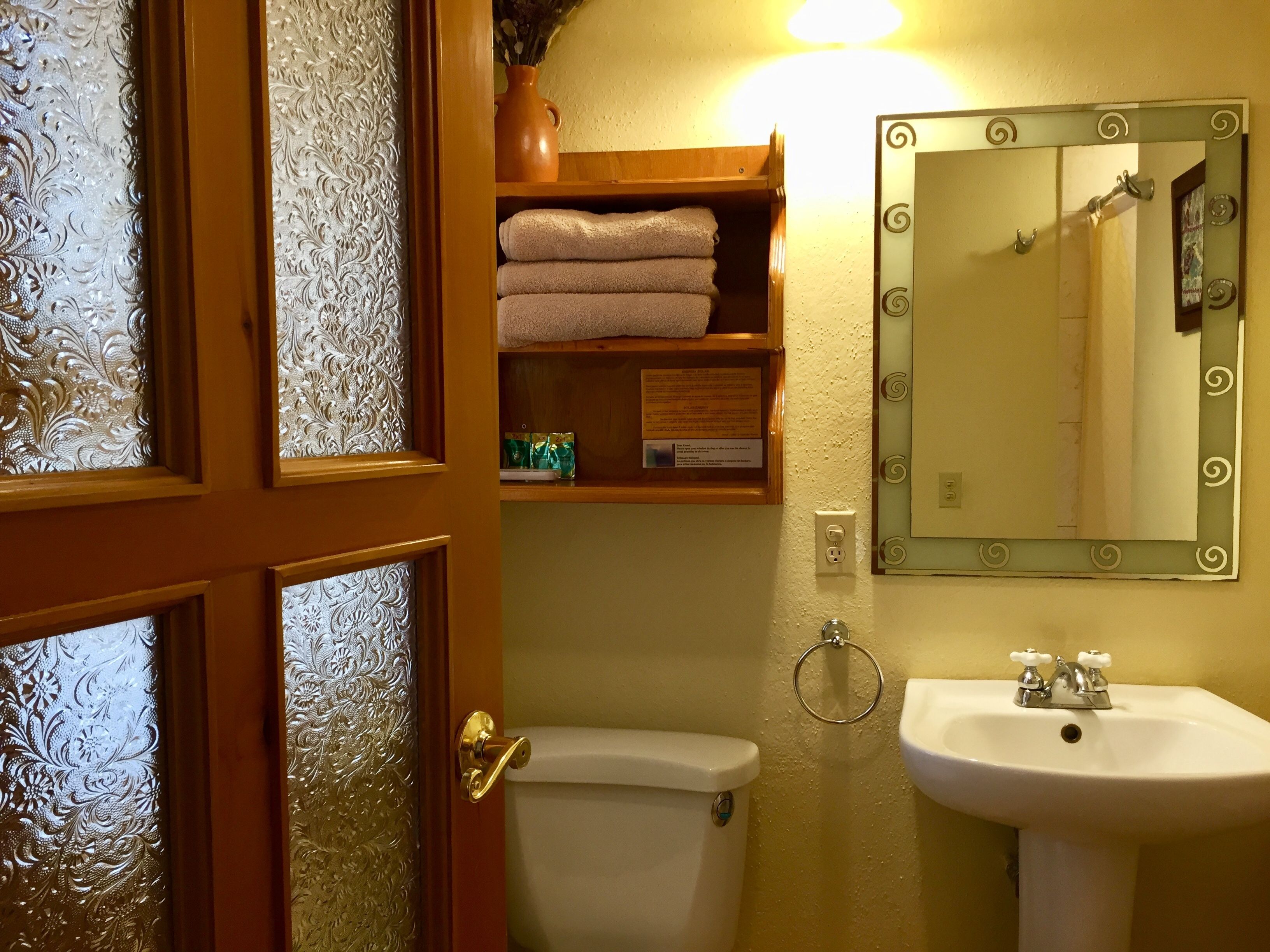 A bathroom with a toilet, sink, and mirror. There is a shelf with towels and a vase with flowers.