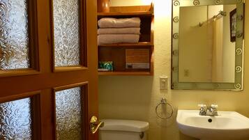 A bathroom with a toilet, sink, and mirror. There is a shelf with towels and a vase with flowers.