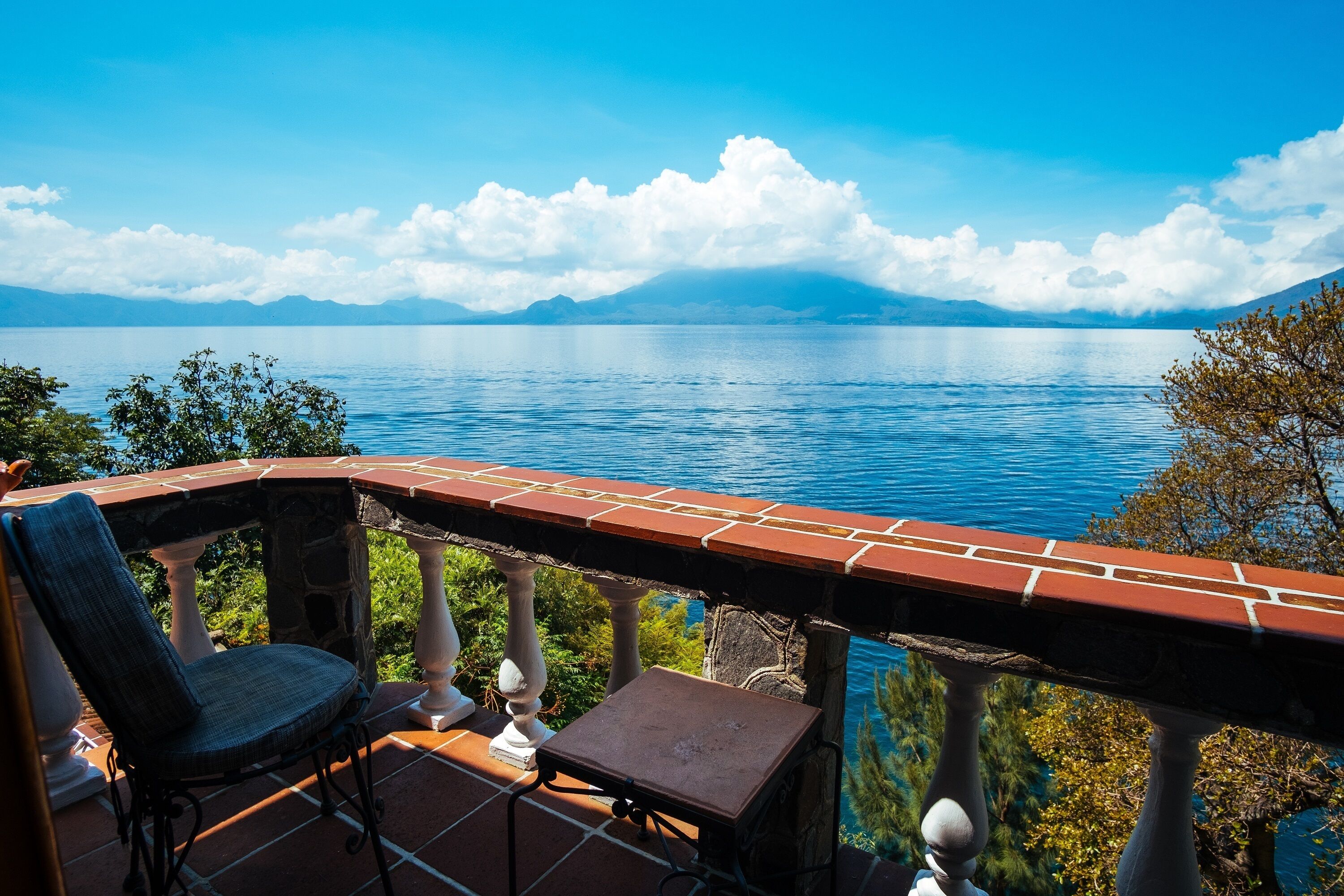 A terrace with a view of a lake and mountains, a chair, and a small table.