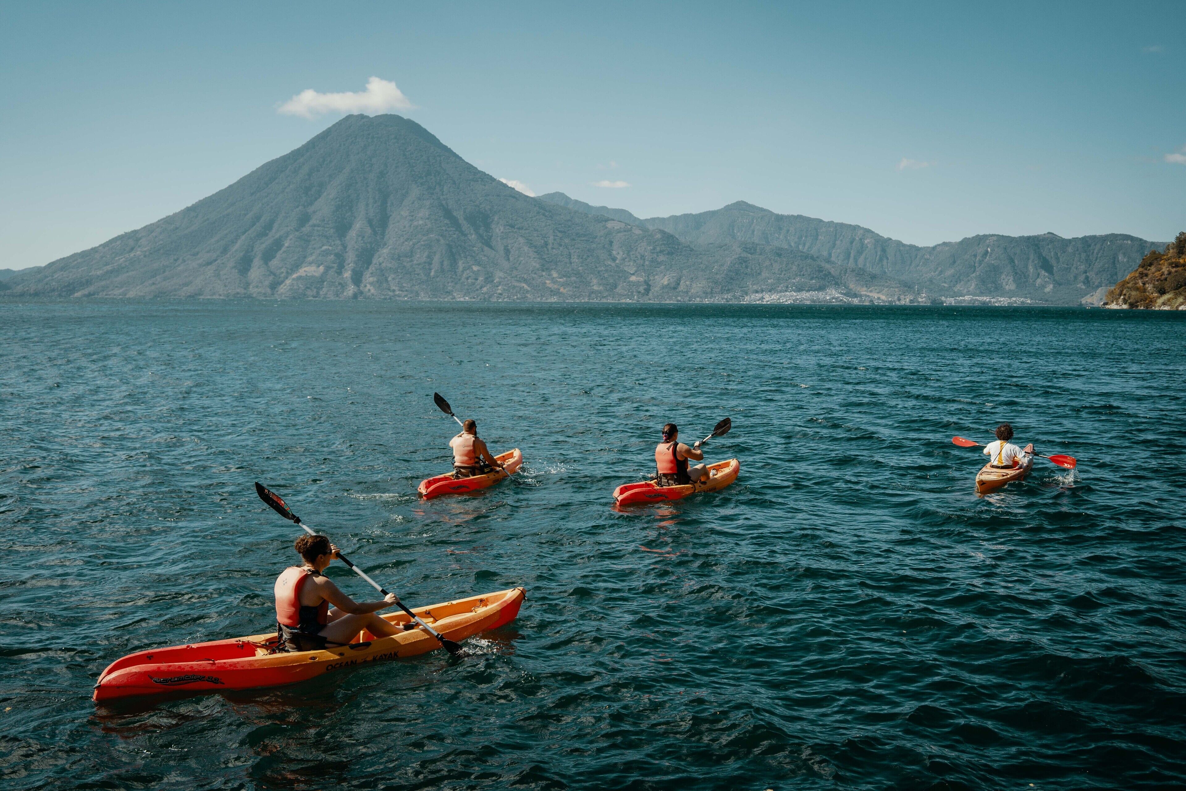 Four kayakers in a row on calm water with a mountain in the background.