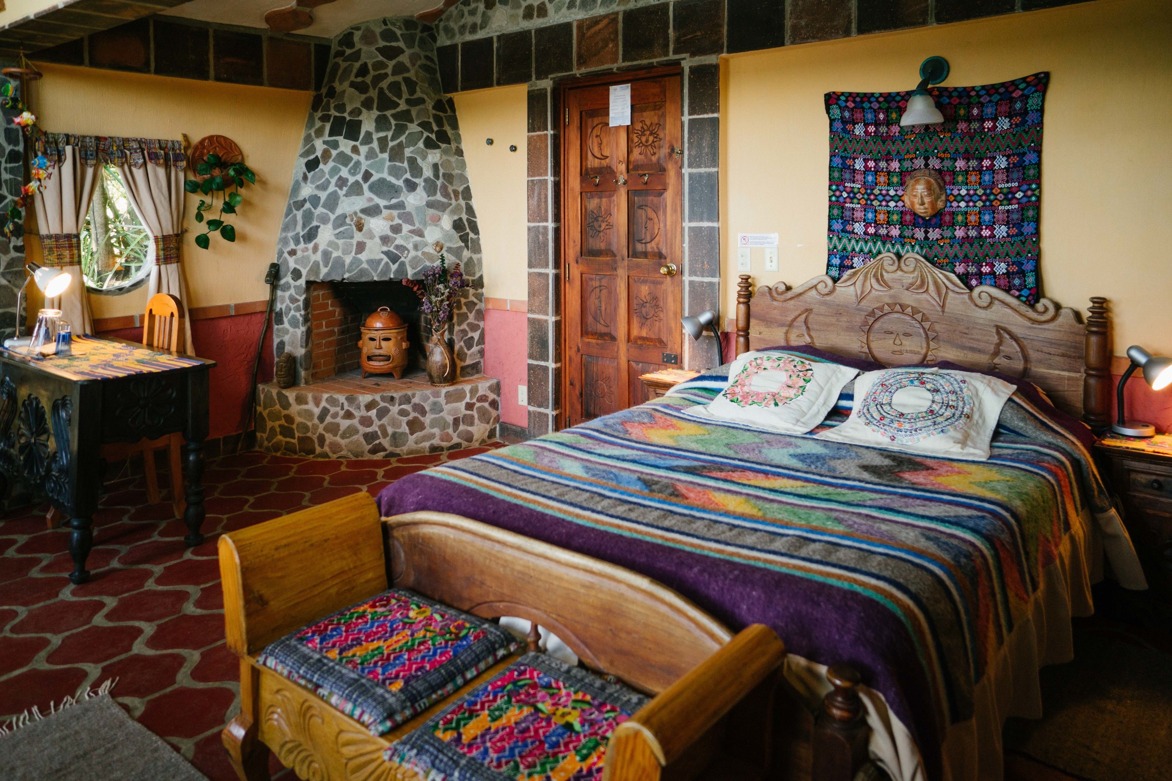 A cozy room with a stone fireplace, a bed with a colorful quilt, a wooden headboard, and a small table with a lamp.