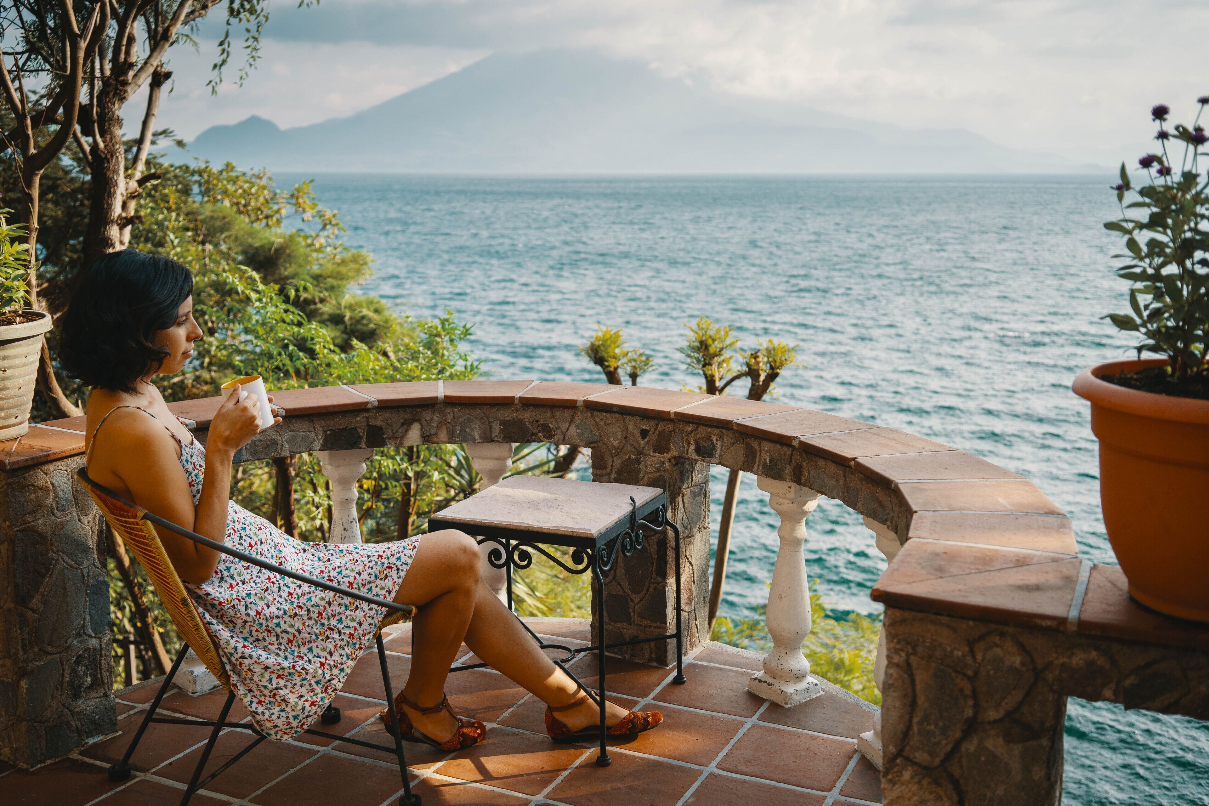 Una mujer sentada en una silla junto a una barandilla de piedra con vistas a un cuerpo de agua y montañas a lo lejos.