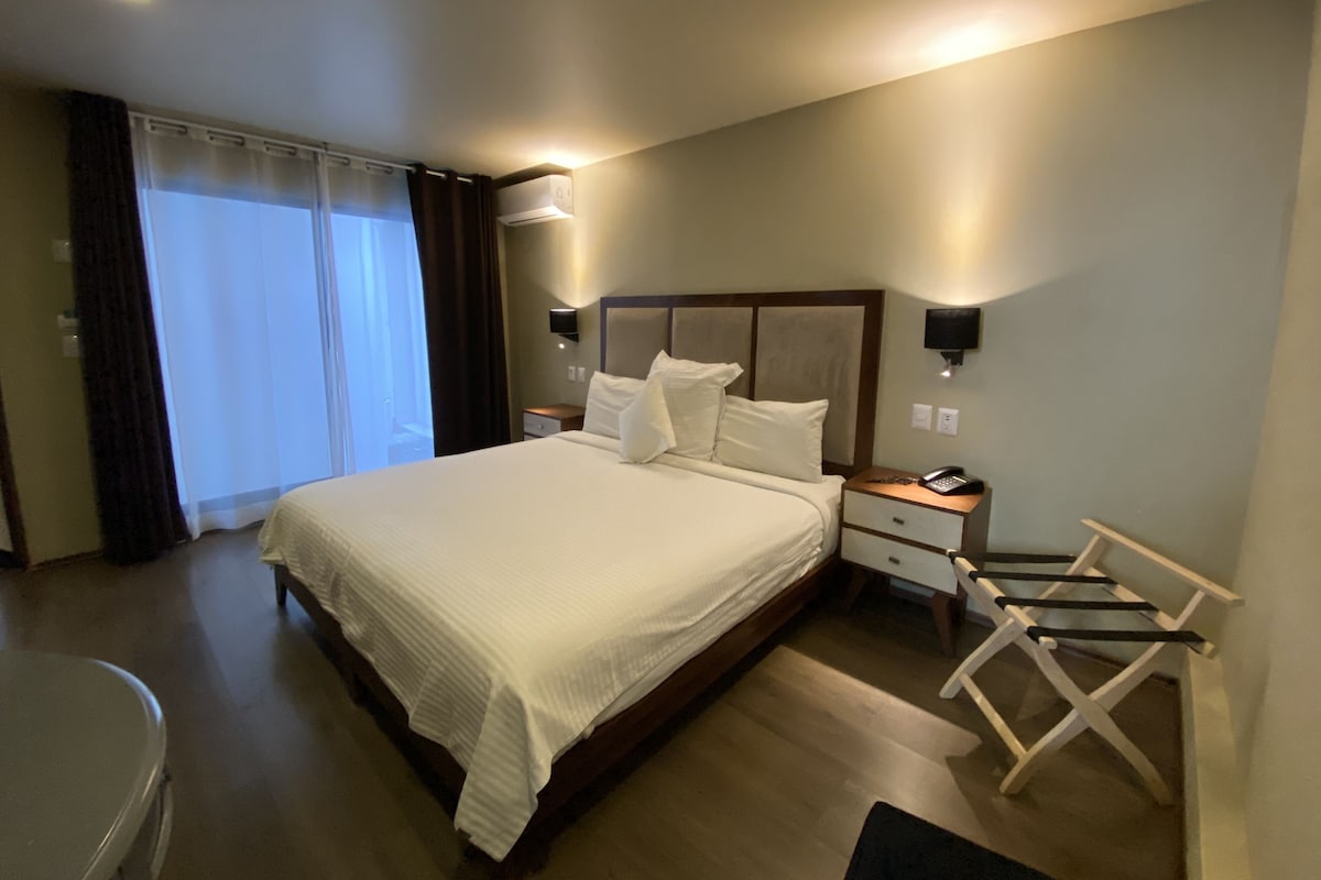 A hotel room with a large bed, a bedside table with a phone, a wall-mounted TV, and a seating area with a chair.