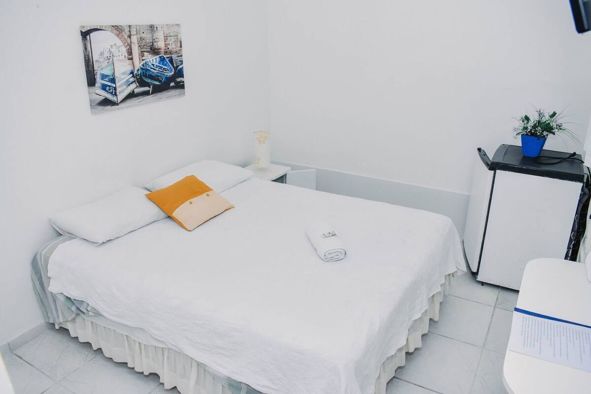 A neatly arranged hotel room with a bed, a small bedside table with a lamp, a potted plant, and a wall-mounted picture.