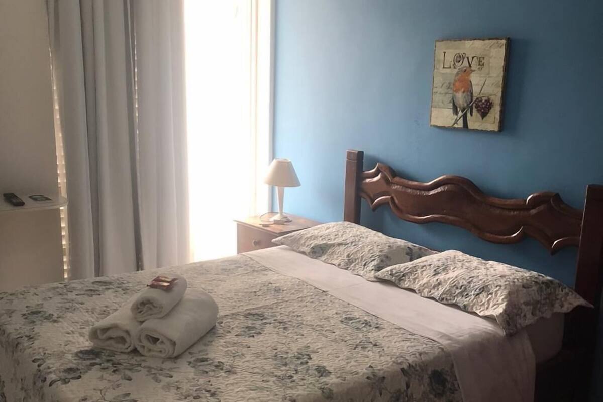 A bedroom with a floral bedspread, a wooden headboard, a nightstand with a lamp, and a wall art piece.