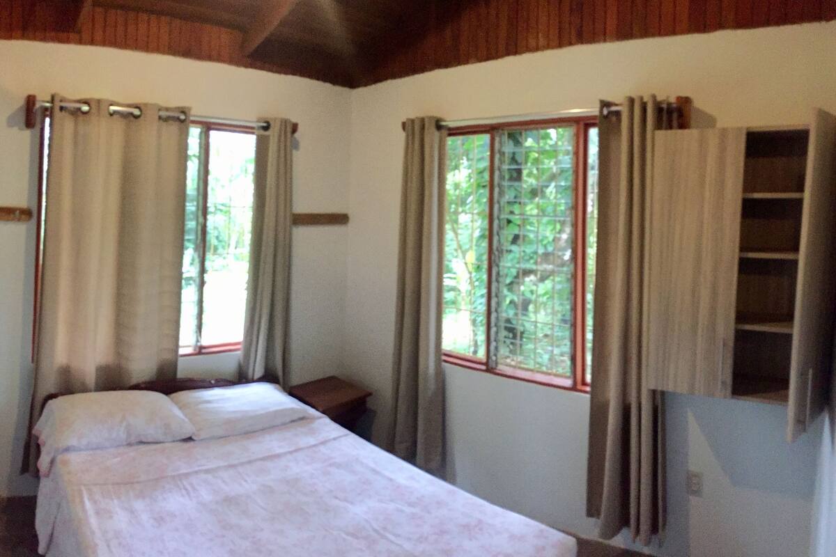 A bedroom with a bed, two windows with curtains, a wooden ceiling, and a bookshelf.