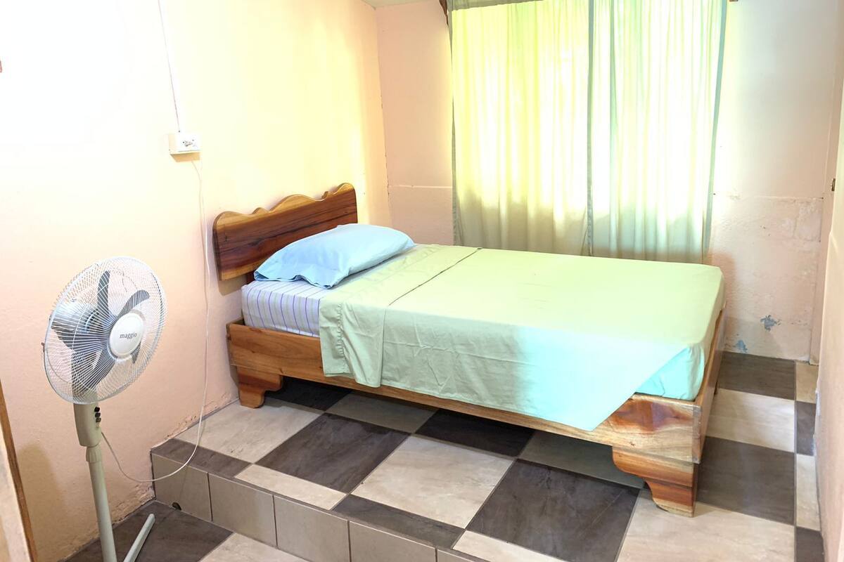 A small, single-bed room with a wooden headboard, a green bedspread, and a checkered floor. A standing fan is visible on the left side.