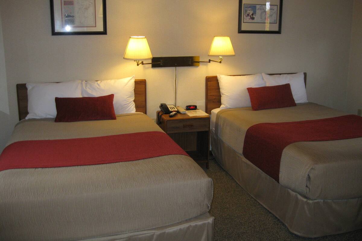 Two double beds with red throw pillows, a nightstand with a phone and alarm clock, and two wall-mounted lamps.