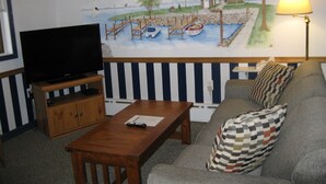 A living room with a television on a wooden stand, a wooden coffee table, a sofa with patterned cushions, and a mural of a coastal scene with a lighthouse, boats, and trees. - Boyne City Motel (Boyne City)