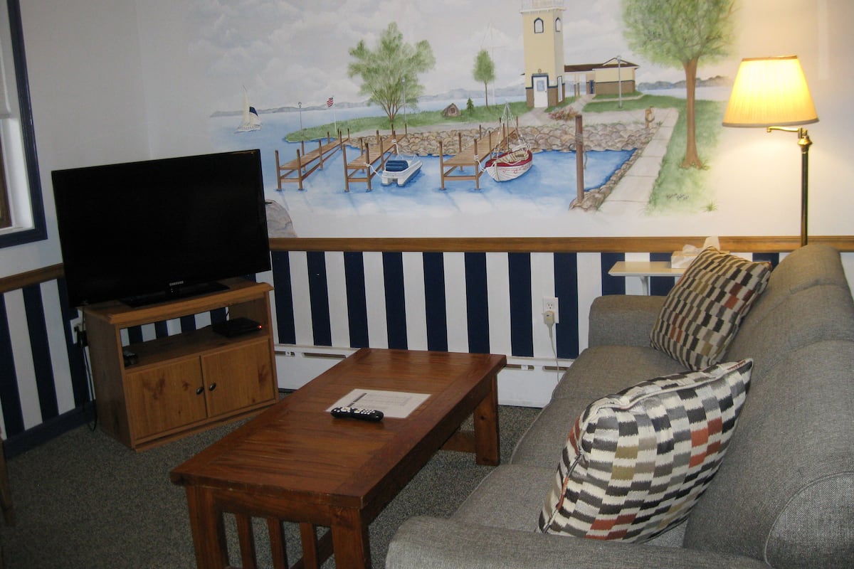 A living room with a television on a wooden stand, a wooden coffee table, a sofa with patterned cushions, and a mural of a coastal scene with a lighthouse, boats, and trees.