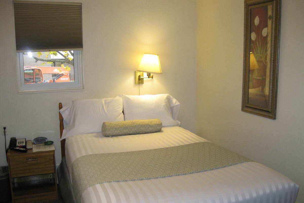 A neatly made bed with a patterned blanket, a bedside lamp, a nightstand with a phone and remote, and a window with blinds.
