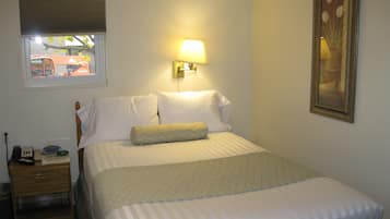 Una cama bien hecha con un edredón estampado, una lámpara de noche, una mesita de noche con un teléfono y un control remoto, y una ventana con persianas.