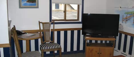 Ein Zimmer mit einem Holztisch, einem gestreiften Stuhl, einem Fernseher auf einem Schrank und einem Fenster mit Jalousien.