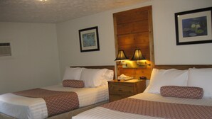 A hotel room with two beds, each with patterned bedding and pillows, a wooden headboard, bedside lamps, and a nightstand with a clock. - Boyne City Motel (Boyne City)