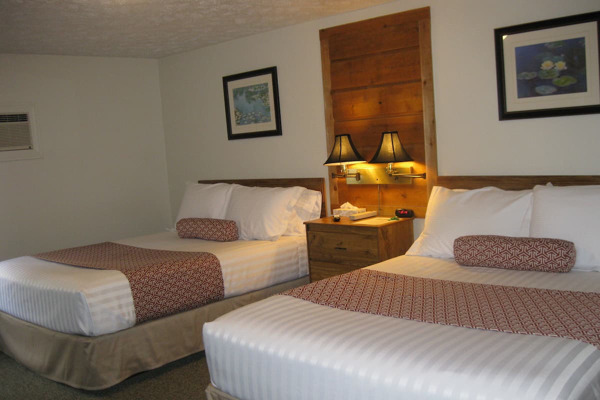 A hotel room with two beds, each with patterned bedding and pillows, a wooden headboard, bedside lamps, and a nightstand with a clock.
