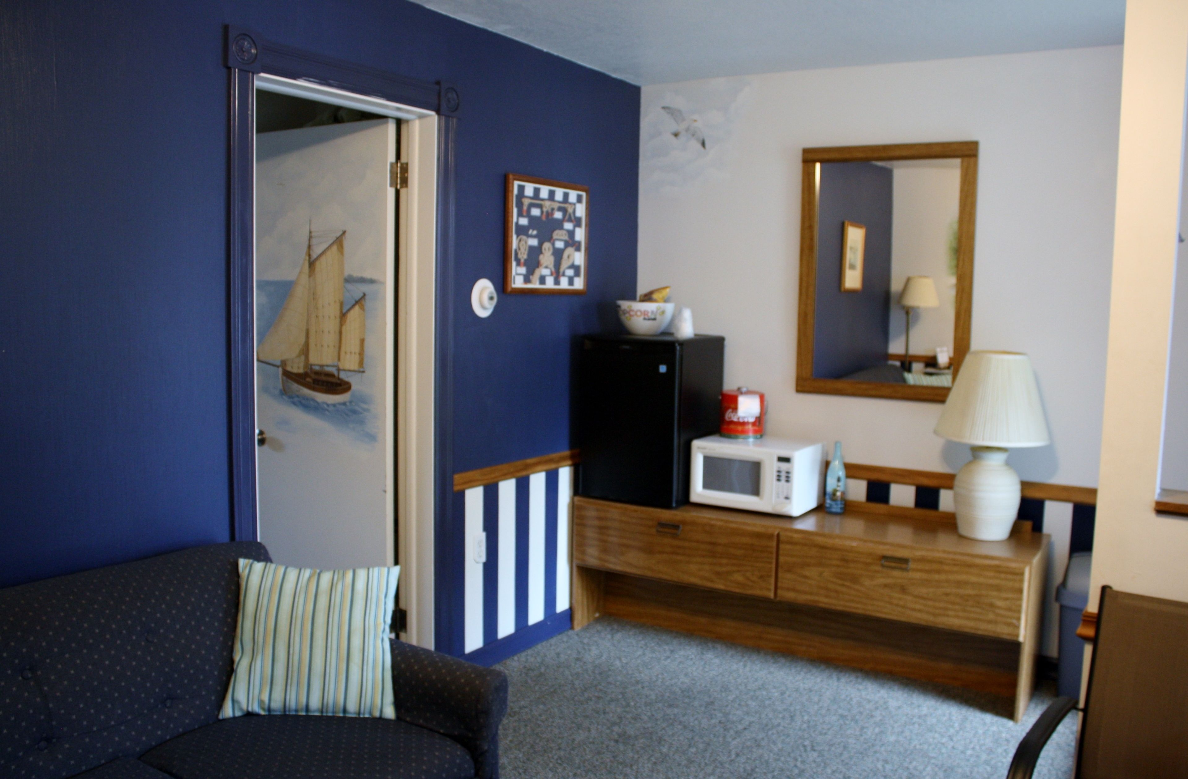 A room with a dark blue wall, a wooden dresser, a microwave, a lamp, a framed picture, and a door with a sailboat painting.