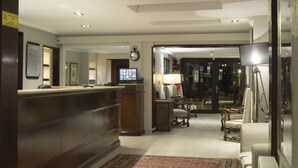 Ein Hotel-Empfangsbereich mit einem Holzbüro, einer Uhr und einem Fernseher. Im Hintergrund stehen Stühle und eine Lampe.