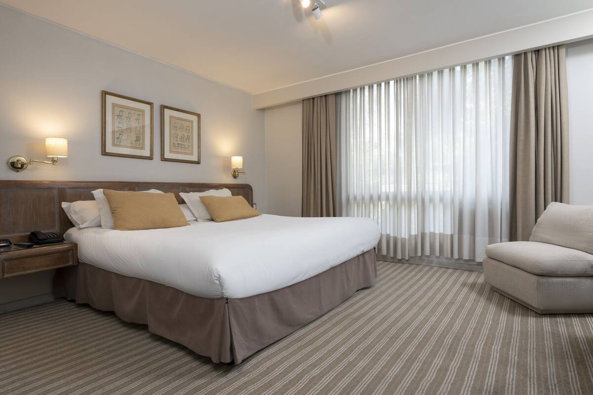Ein Hotelzimmer mit einem großen Bett, einem Nachttisch mit Lampe, einem Sessel und einem großen Fenster mit Vorhängen.