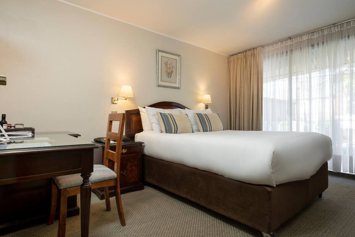 Ein Hotelzimmer mit Bett, Schreibtisch, Stuhl, Lampe und einem Vorhangfenster.