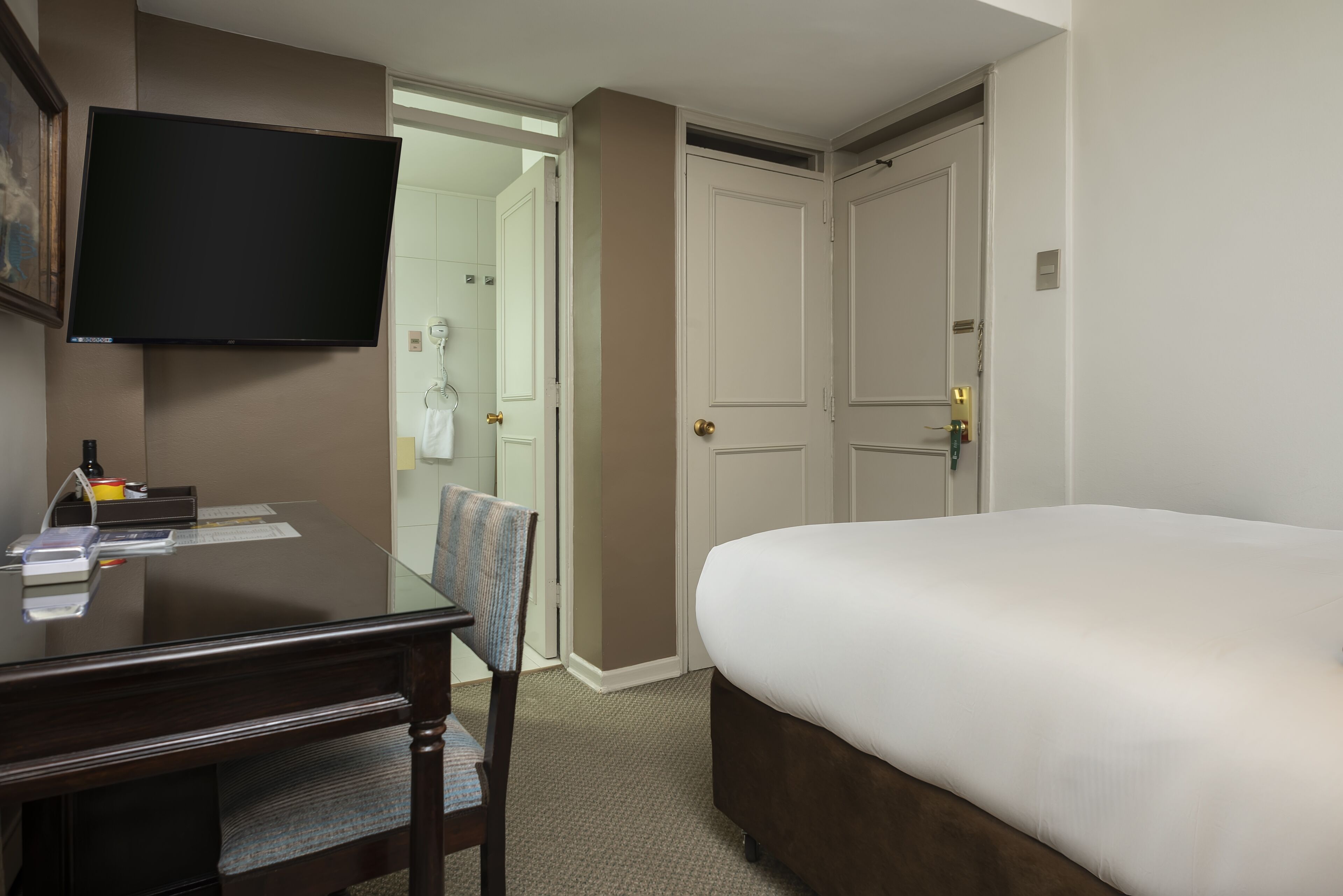 A hotel room with a bed, a desk with a chair, a flat-screen TV mounted on the wall, and a door leading to a bathroom.